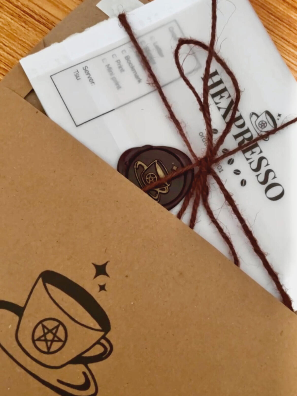 the hexpresso letter club envelope close up details kraft envelope with a the Hexpresso coffee cup logo. The cup has a pentacle and two four pointed star sparkles above. Envelope contents peak out, wrapped in vellum paper with the Hexpresso name and logo. Dark brown wax sealed with Hexpresso logo and wrappe